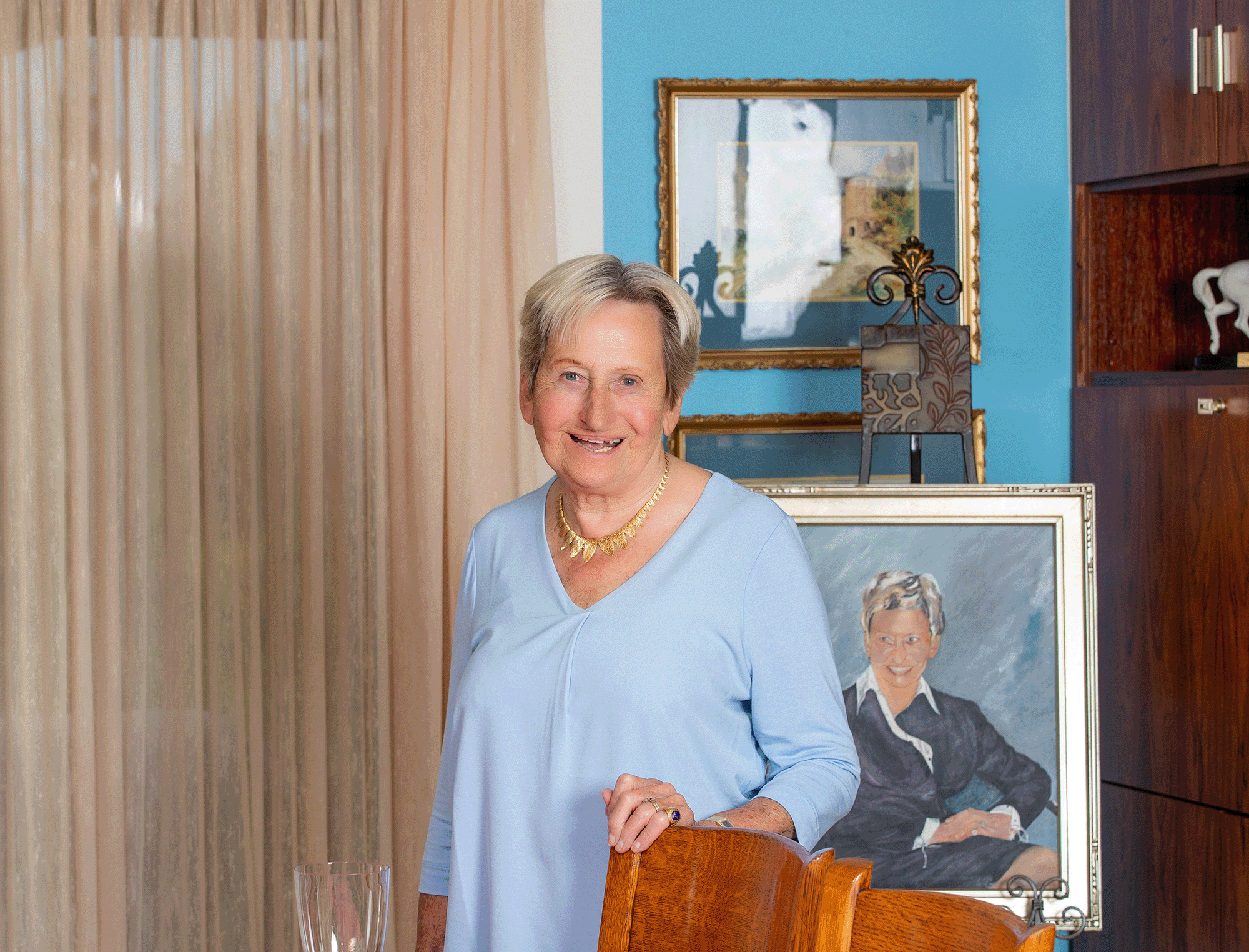 A photo of Margaret Juravinski. She has her hand on the top of a chair and is standing in front of a painted portrait of herself. 